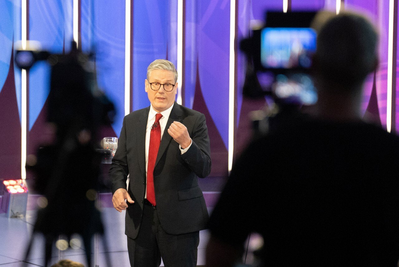 A man in a suit speaking, seen from behind two television cameras.